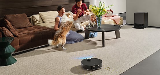 Robot sprzątający odkurza dywan w salonie, obok siedzi rodzina z dzieckiem i psem